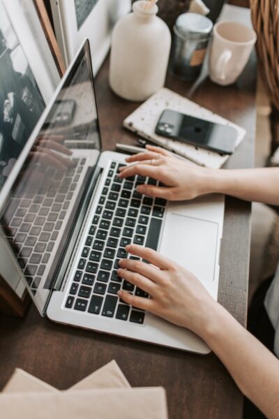 From above of unrecognizable woman sitting at table and typing on keyboard of computer during remote work in modern workspace